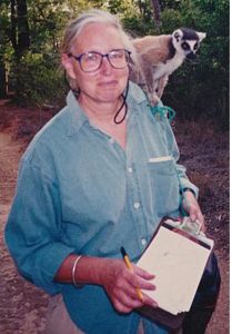 Alison Jolly shown undertaking field work, holding a clipboard and pen and looking at the camera. A ring-tailed lemur perches on her left shoulder.