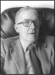 Face and torso of an elderly Donald Hebb, wearing spectacles, suit and tie, sitting in an armchair