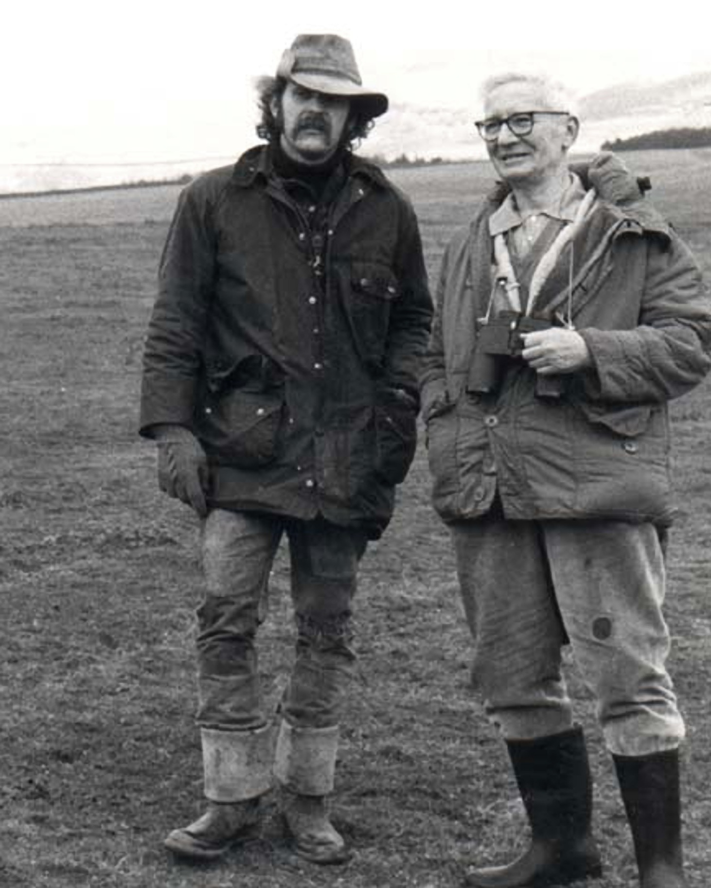 Tinbergen, wearing outdoor clothes and with binoculars around his neck, is standing in a rural landscape with a companion