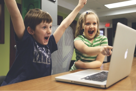 Your gurl and boy on laptop, Girl in green stripey top pointing at screen, boy in blue T-shirt has both arms in the air celebrating. They appear to be in a computer lab