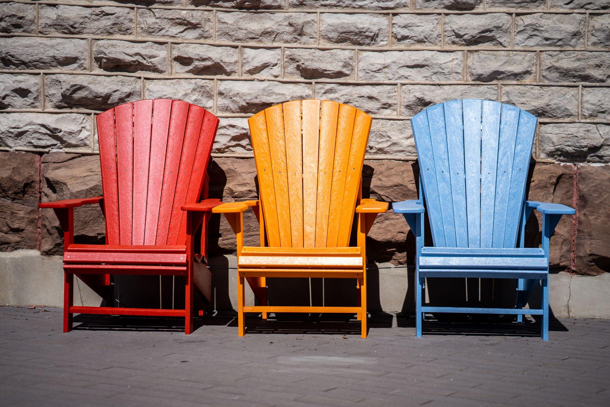 Three Wooden outdoor garden chairs, one red, one orange and one blue, sitting on black brick floor with a sandstone wall behind them