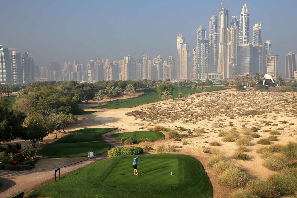 Photograph of a golf course in Dubai with several turfed golf greens and someone playing golf, set within an arid landscape and against a distant cityscape including tall buildings