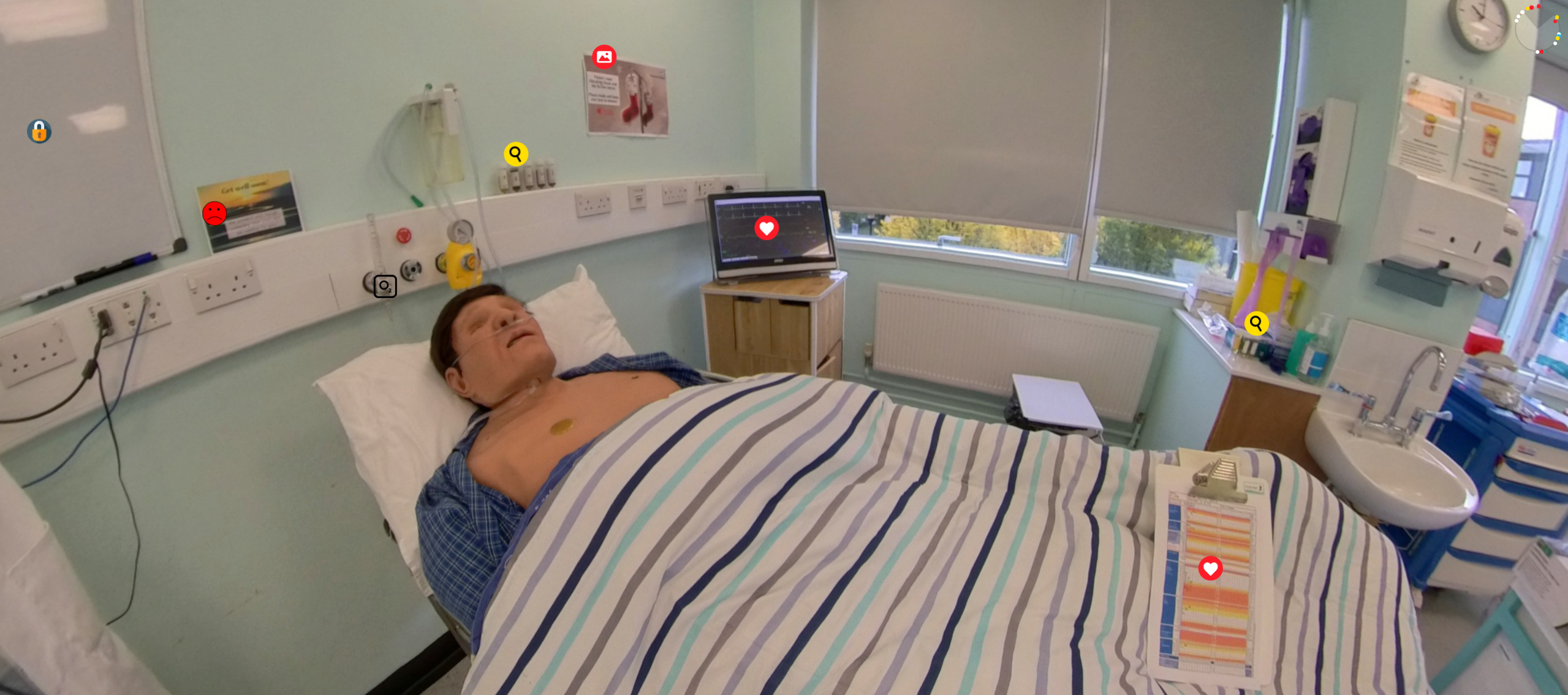 A 360-degree view of a simulated hospital room with multiple beds. The room has a light blue colour scheme and is well-lit with ceiling lights. In the centre, a mannequin lies in one of the hospital beds, covered with a striped blanket. The room is equipped with medical devices, monitors, and various medical supplies on trolleys. There are also a couple of cribs and an open window that allows natural light to enter. The environment appears clean and organised, typical of a healthcare facility.