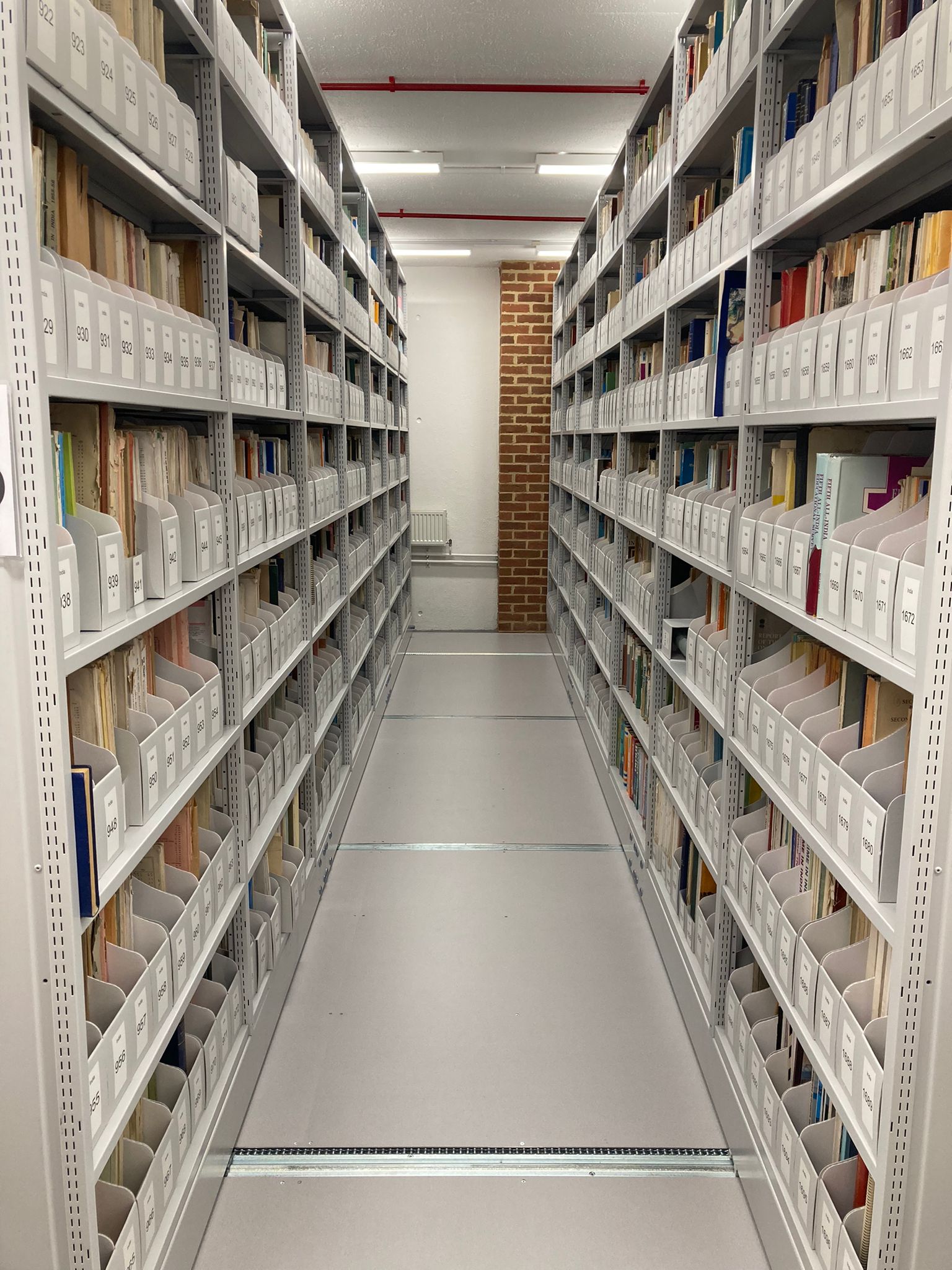 Image credit: Photograph of rolling stacks in the BLDS Legacy Collection. Copyright University of Sussex (2024). Used with kind permission. All rights reserved. Photograph taken in an archive of a corridor with shelves on either side. On each shelf are documents and these represent the BLDS Legacy Collection.