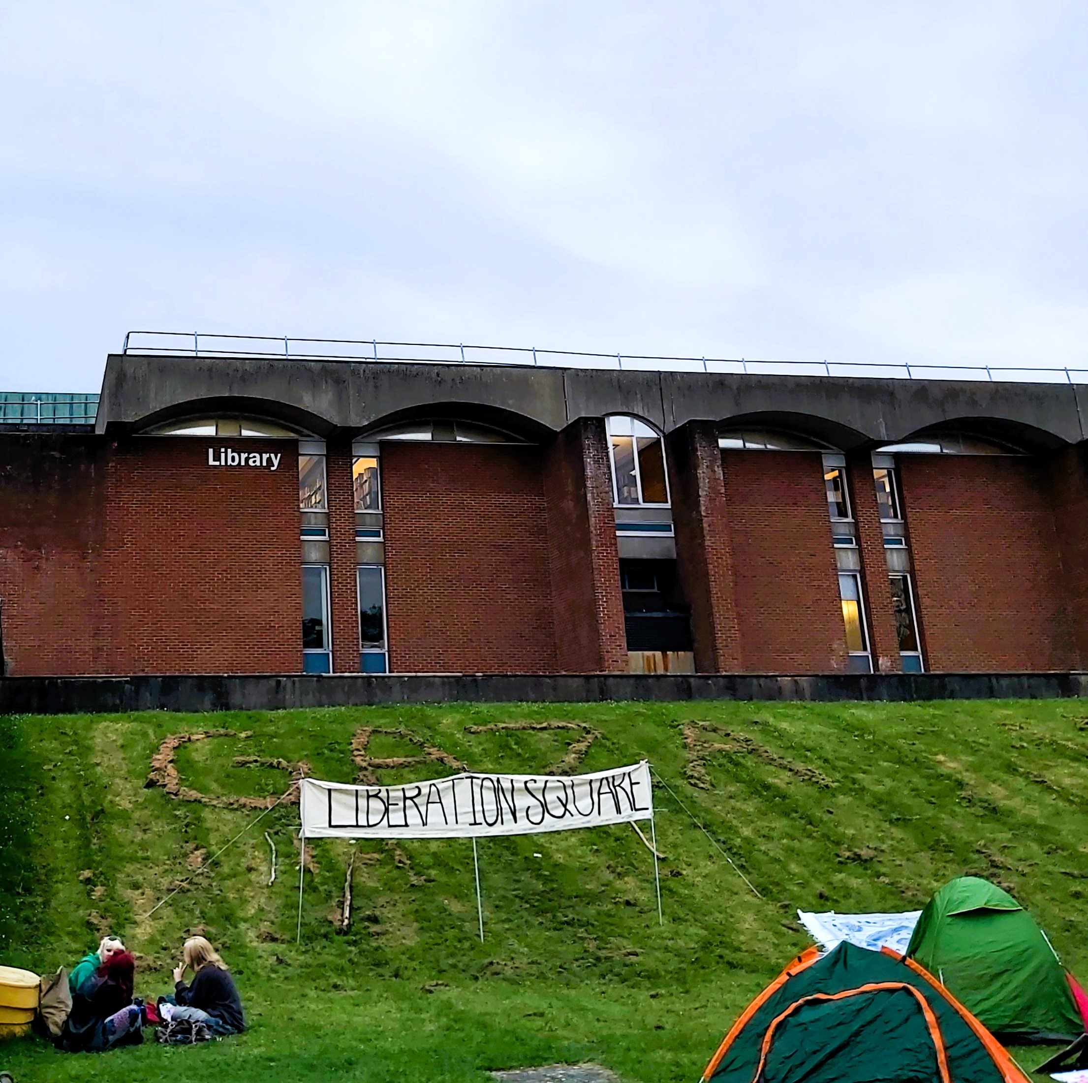 Image credit: Photo copyright Alice Corble (2024). Palestine liberation encampment outside University of Sussex Library in Library Square. Under a CC BY 4.0 licence: https://creativecommons.org/licenses/by/4.0/deed.en Exterior of the front elevation of University of Sussex Library, a redbrick building in a Modernist style wit the word Library on the wall. The building is on top of a freshly mown sloping lawn and the word GAZA has been spelled out in large capital letters on the bank with the grass cuttings. Beneath this at the bottom of the slope is a large hand-made cloth banner reading LIBERATION SQUARE, with students sitting cross legged on the lawn on the left and dome shaped tents on the right.
