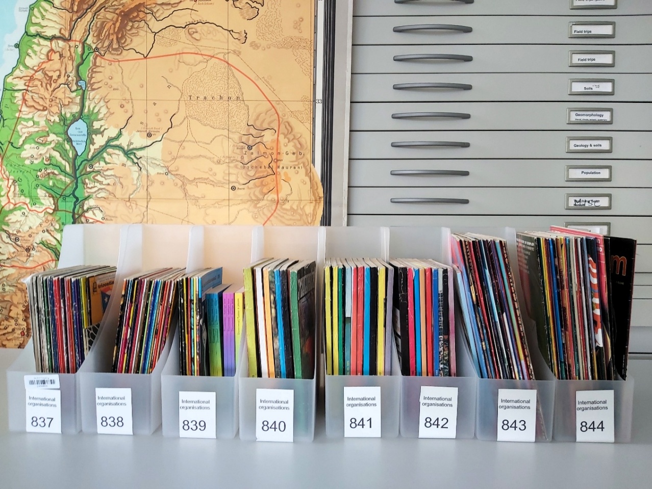 Image credit: OSPAAAL periodicals in archive boxes from the BLDS Legacy Collection workshop, University of Sussex. Photograph copyright Alice Corble (2024). Licence: CC BY 4.0. https://creativecommons.org/licenses/by/4.0/deed.en Eight numbered archival boxes containing periodicals with colourful spines lined up on a table. Behind them on the wall is a large format map and a planchest of map drawers.