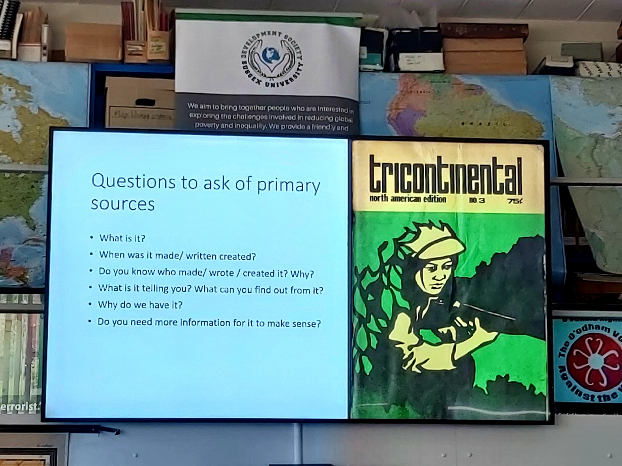 Image credit: 'Questions to ask of primary sources'. Slide by Danny Millum (2024). Photograph by Alice Corble. Photograph of a large LED screen against a backdrop of maps and boxes in a workshop room. The screen displays a teaching slide by Danny Millum. The text on the slide reads: Questions to ask of primary sources: What is it? When was it made/written/created? Do you know who made/wrote/created it? Why? What is it telling you? What can you find out from it? Why do we have it? Do you need more information for it to make sense? The image on the screen to the right of these questions is the yellow, green and black cover of Tricontinental Magazine, North American Edition no 3. The cover image is a soldier in a bush pointing a rifle. Photograph by Alice Corble.