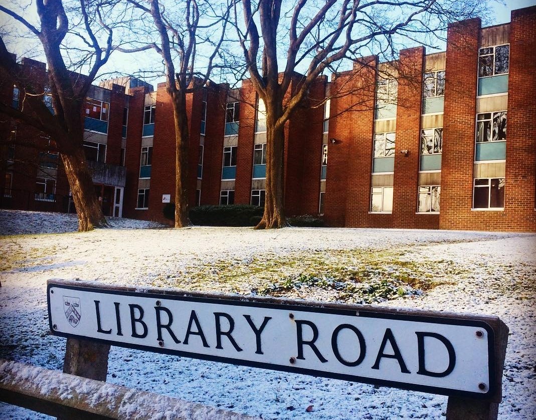 Credit: Photograph by Carolsmithyes CC BY-SA 4.0, https://commons.wikimedia.org/w/index.php?curid=73859036 Front elevation of the Institute of Development Studies (IDS) redbrick building in winter. In the mid-ground is three leafless trees on a white frost-covered lawn. In the foreground is a kerbside white road sign with LIBRARY ROAD inscribed in black capital letters next to a University of Sussex coat of arms emblem.