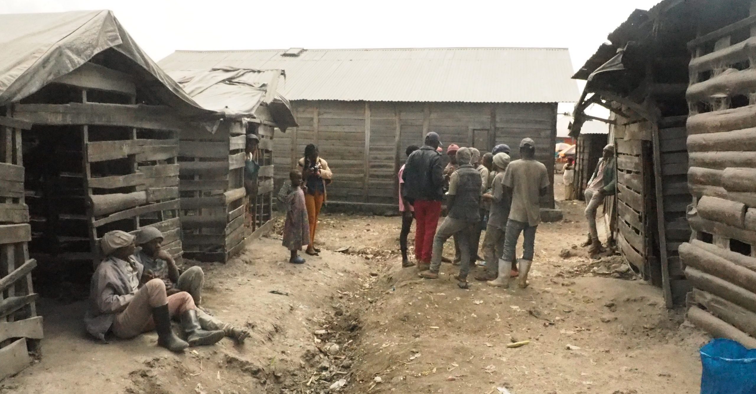 A photo of people waiting for work in Kitchanga. There is a group of people standing in between wooden houses.