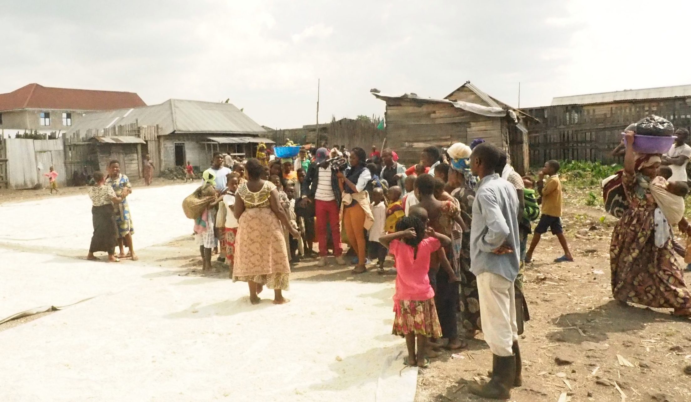 A photo showing the filming of Migrations. There is a group of adults and children gathered together with wooden houses in the background. In the centre of the group, a woman is holding a film camera.