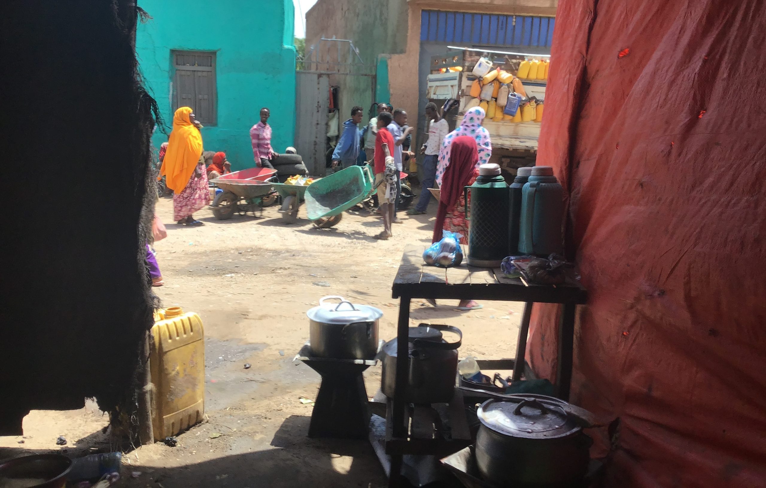 A photo of a busy street. In the foreground there are cooking pots and in the background there are people pushing wheelbarrows with water containers behind them.