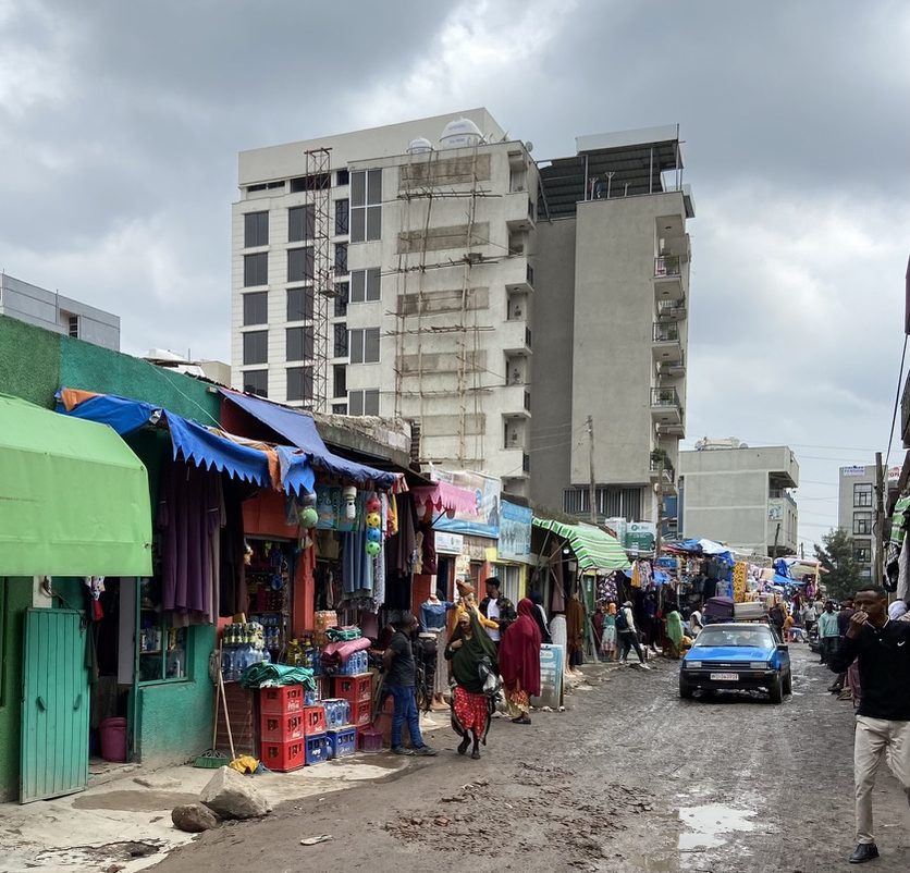 A photo of an urban scene. In the foreground is a muddy street with colourful shopfronts along the sides. A car is driving down the road and people are walking along the sides of the road. In the background is a tall block of flats under construction.