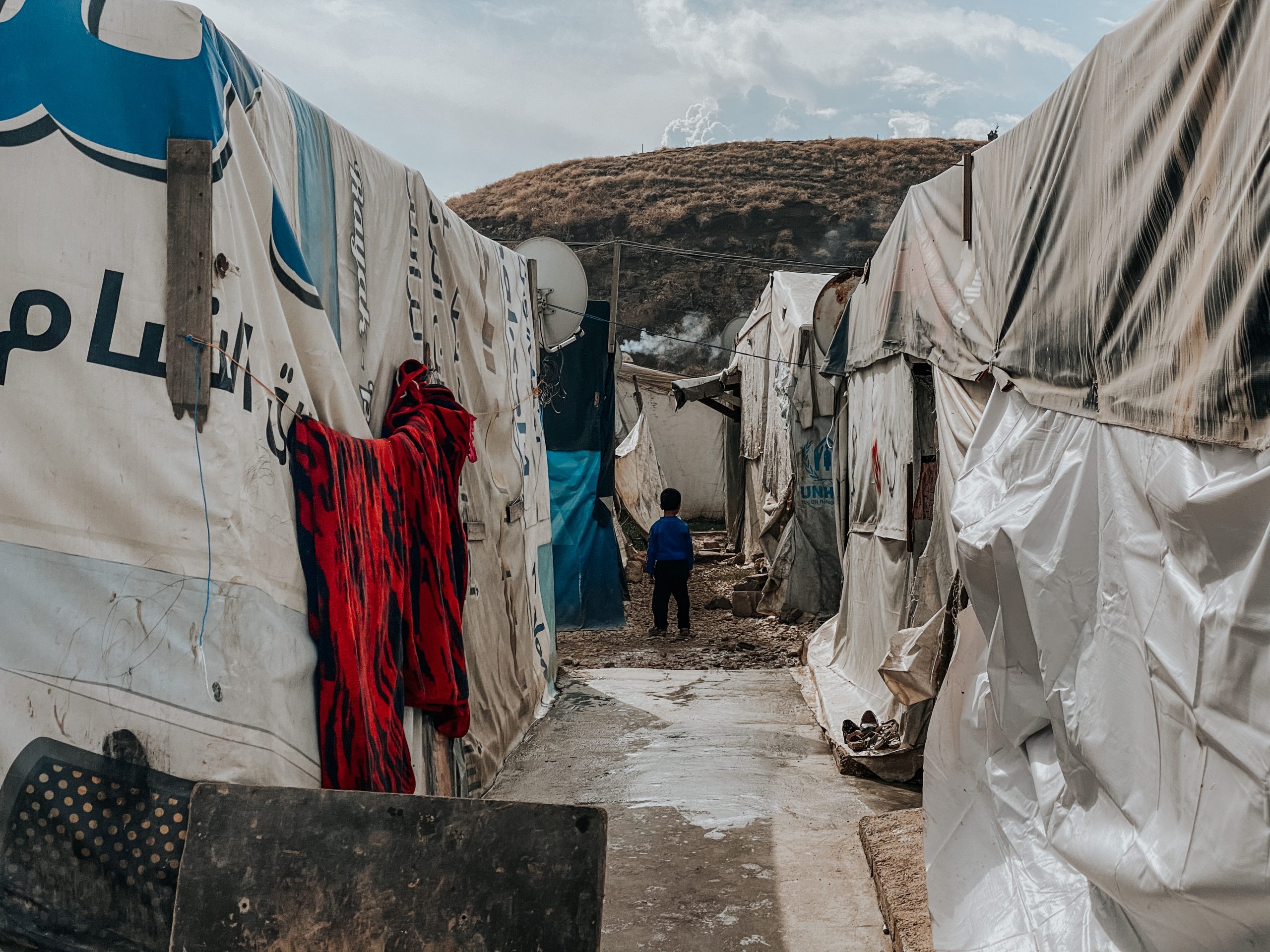 Photo of a refugee camp in Bekaa. The perspective is of a walkway between two rows of tents. A small boy is standing with his back to the camera in the centre of the image. In the distance there is a hillside covered in dry bushes.