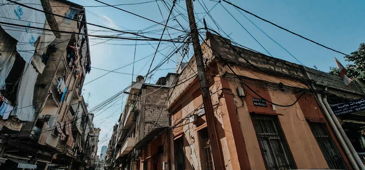 A photo of the corner of a street in an urban setting taken from the ground level and looking upwards. There are buildings and many cables running between them.