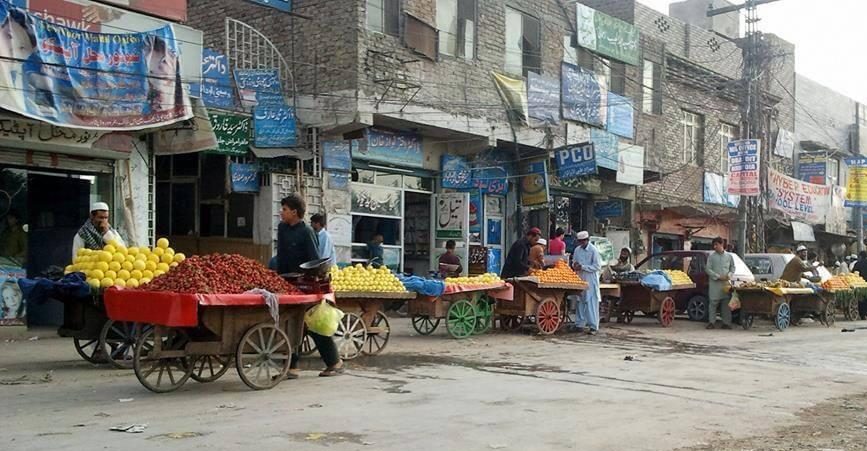 A photo of a street scene showing market stalls and vendors selling fruit and vegetables. Many stalls are on wheeled carts. Behind the stalls are buildings.