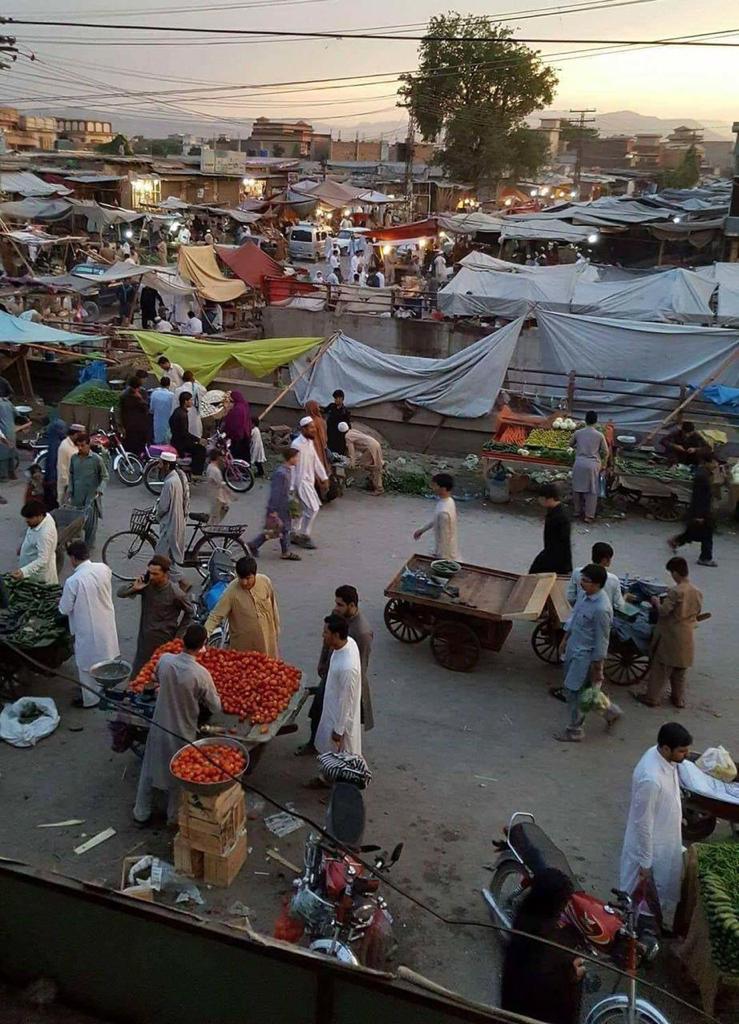 A photo of an urban setting. There are market stalls filled with food with buildings in the distance. There are lots of people in the foreground – many are travelling on bikes. Overhead cables criss-cross the sky.