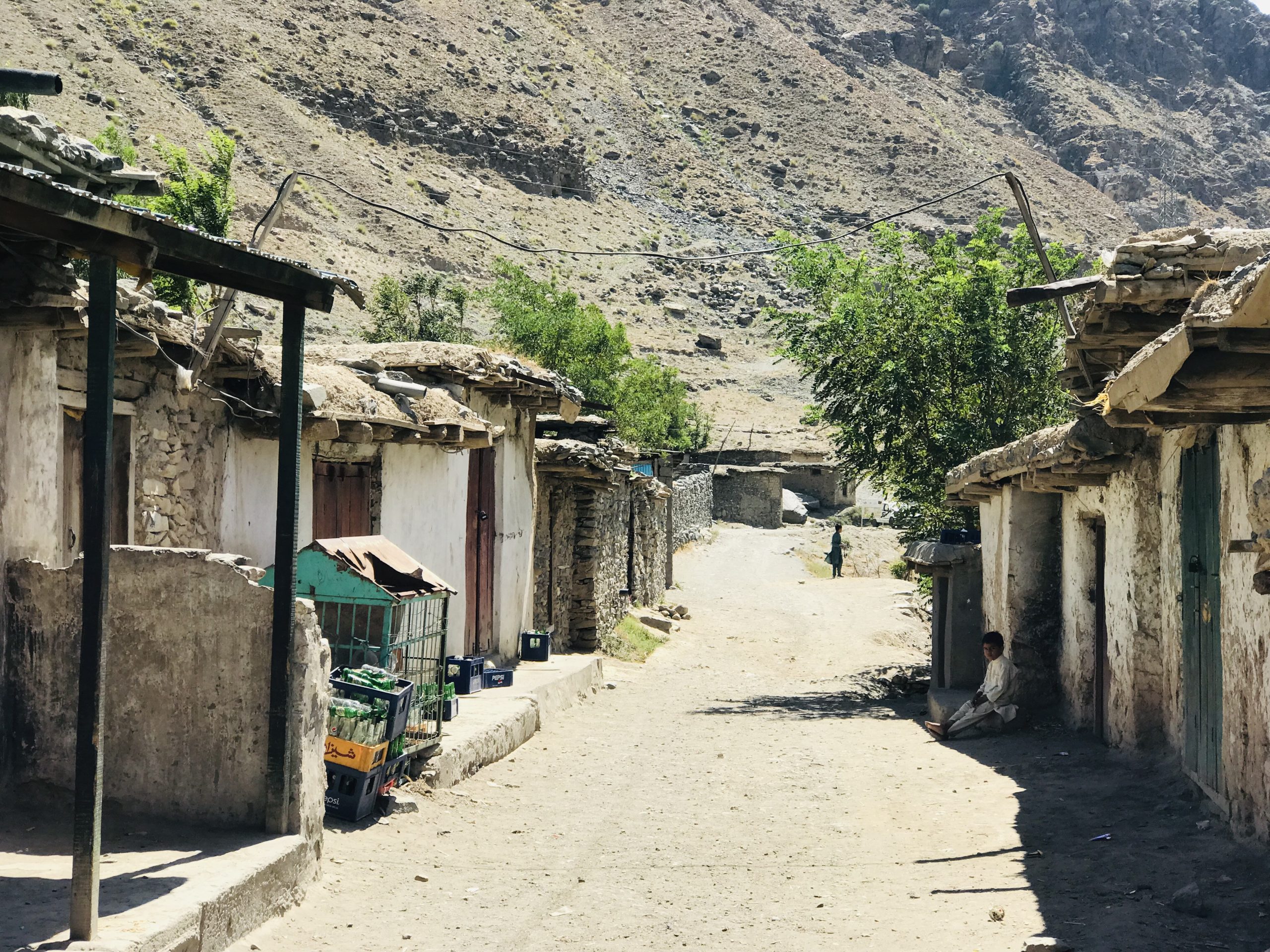 Photo of a refugee camp. The image is taken from ground level in a street with houses on both sides. A small boy is visible in the distance, otherwise there are no other people. There are mountains in the background.