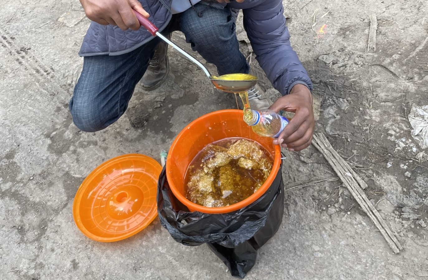 A photo of a person pouring honey from an orange bucket into a plastic bottle with a ladle.