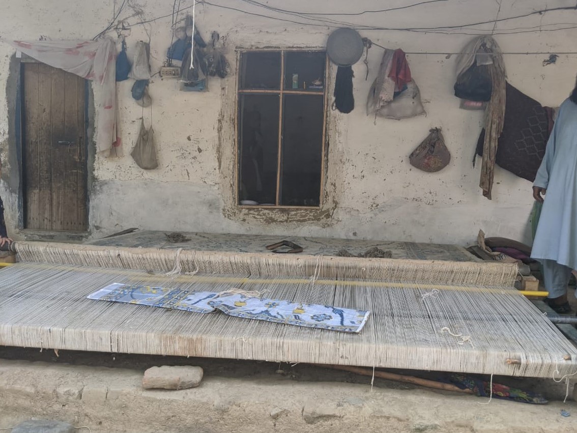 Photo of a loom with a partially finished carpet on it. It is in front of a house.