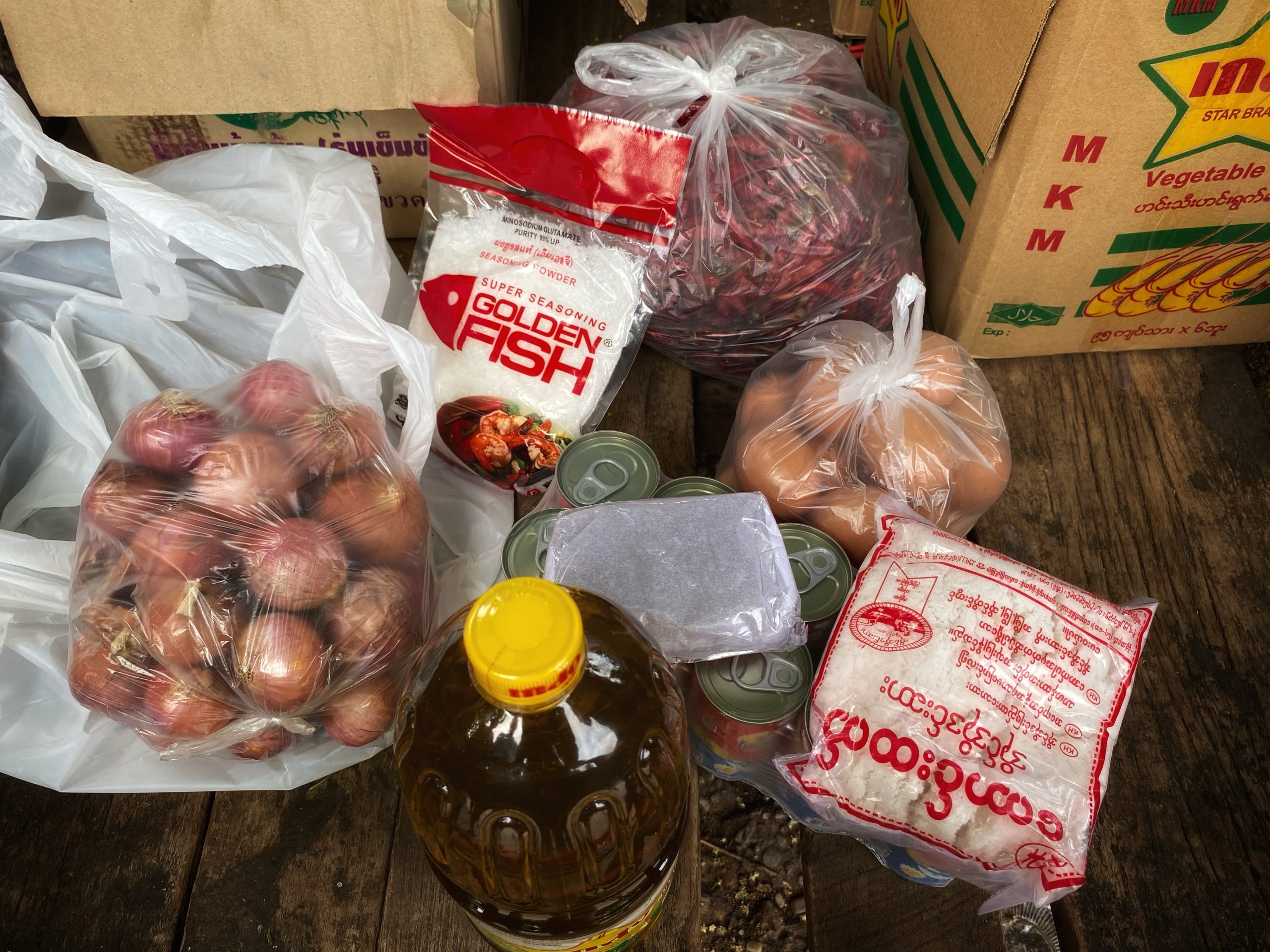 Photo of food items including a bottle of cooking oil, bags of onions and eggs, packets of rice, and tins.