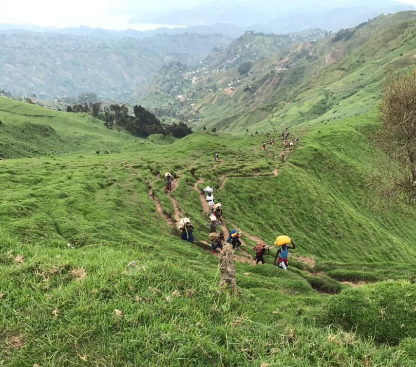 Photo of people walking through the countryside. There are green, grass-covered hills and people walking through them along tracks carrying packs and water containers on their shoulders.