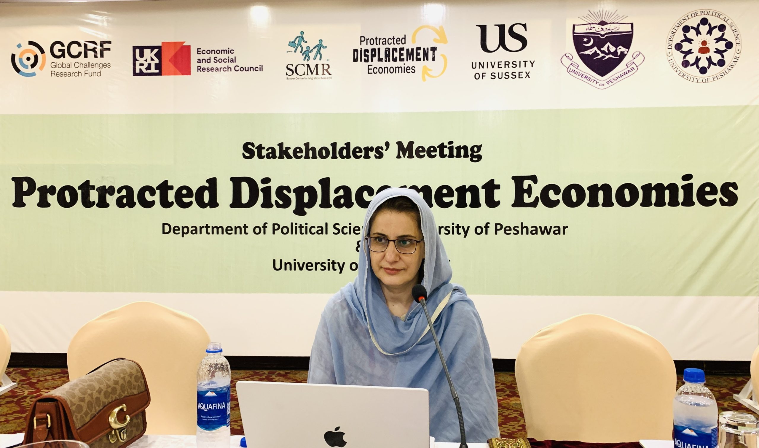 A photo of a woman sitting behind a microphone with a laptop on the table in front of her. Behind her is a banner that reads ‘Protracted Displacement Economies’.