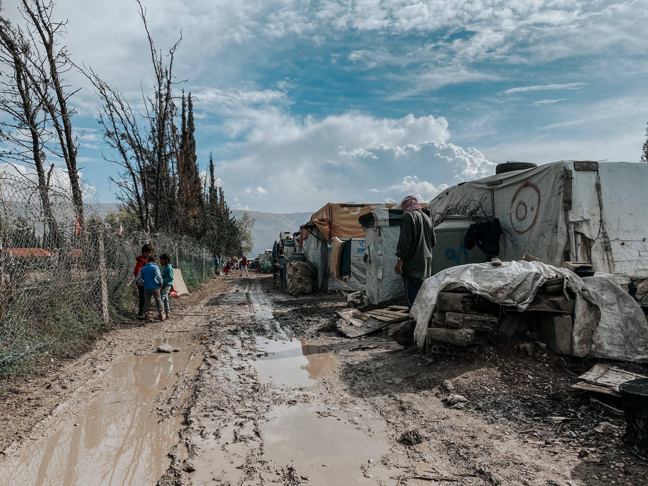Photo showing a refugee camp. On the left-hand side, children are gathered next to a fence with barbed wire along the top. In the middle is a muddy track. On the right-hand side there are adults standing outside tents.