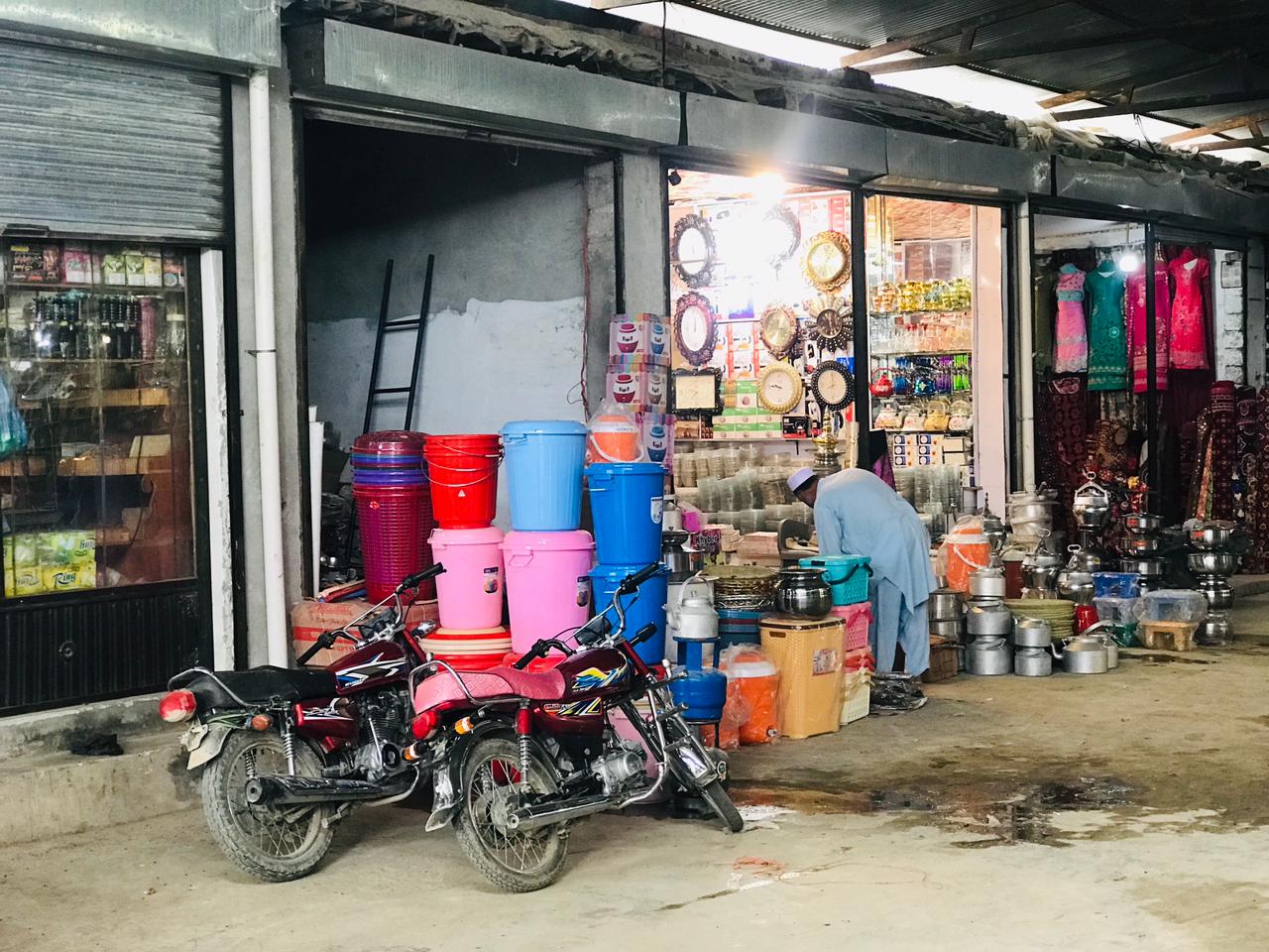 A photo of an urban street with shops selling household goods such as buckets and pans, as well as clothing. In the foreground are two motorcycles.